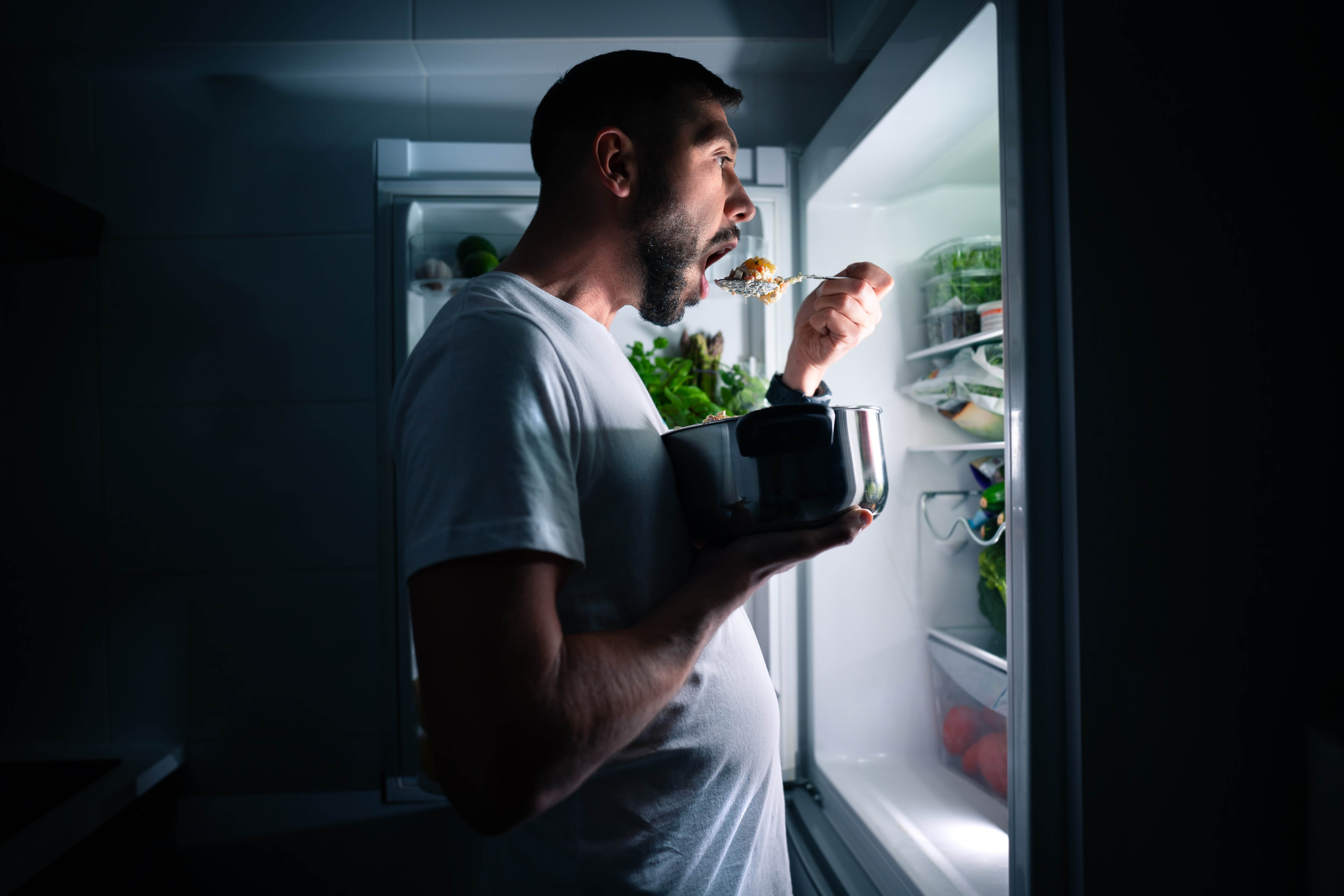 Man standing and eating with the fridge door open and the fridge light on him Man standing and eating with the fridge door open and the fridge light on him